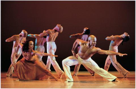 male dancers in white pants walk forward with heads up while a man and a woman in front of them lunge backwards