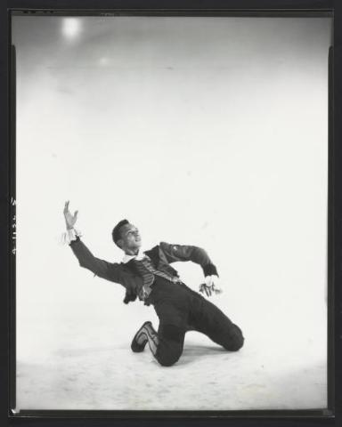 A black-and-white photo of Talley Beatty performing a dance move, captured in a dynamic pose with one knee on the ground and one arm dramatically extended upwards. He is wearing a ruffled shirt and dark pants, set against a plain light background.