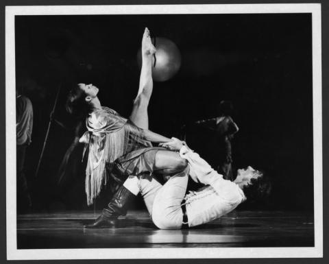 A black-and-white photo of dancers Donna Wood and Peter Woodin performing in Eleo Pomare's Blood Burning Moon. Woodin is lying on the ground while supporting Wood, who has one leg extended high in the air and is dressed in a fringed costume, set against a dark background with other performers faintly visible.
