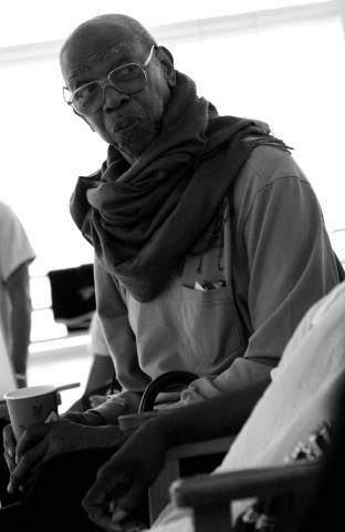 A black-and-white photo of Geoffrey Holder wearing glasses and a scarf, seated and holding a cup. He looks towards the camera with a thoughtful expression, the background showing a light-filled room with blurred figures.