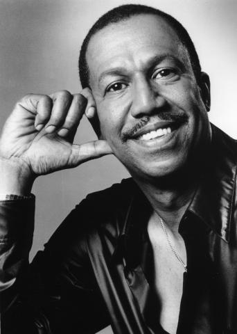 A black-and-white portrait of George Faison, smiling and resting his head on his hand. He is wearing a dark, open-collared shirt, and the background is plain and light-colored.