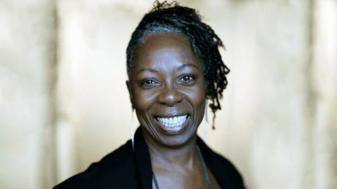 A portrait of Jawole Willa Jo Zollar, smiling broadly. She is wearing a dark top with her hair styled in twists, set against a softly blurred background.