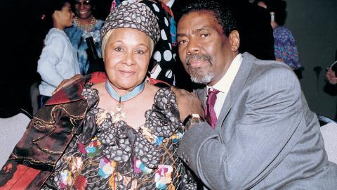 A color photo of Katherine Dunham and Alvin Ailey seated together. Dunham is wearing a patterned dress and headscarf, while Ailey is in a gray suit and red tie; they both smile gently at the camera.