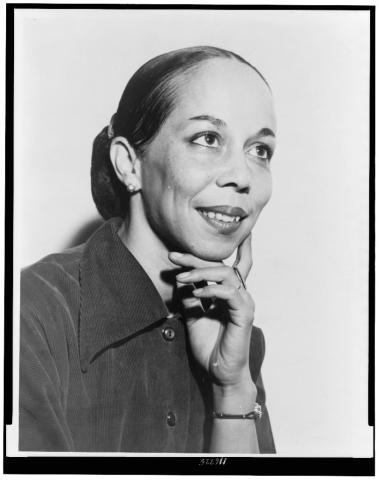 A black-and-white head-and-shoulders portrait of Janet Collins, facing right and resting her chin on her hand. She is smiling and wearing a dark collared blouse, with her hair neatly pulled back.