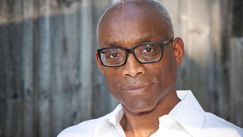 A portrait of Bill T. Jones, wearing a white shirt and black-rimmed glasses. He stands in front of a wooden fence, looking directly at the camera with a composed expression.