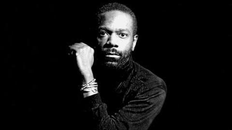A black-and-white portrait of Ulysses Dove, with a focused expression and his arm raised, wearing a dark shirt. The background is completely black, highlighting his intense gaze and strong presence.