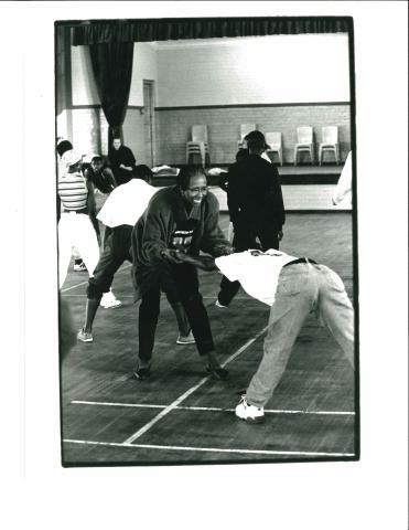 A black-and-white photo captures an energetic moment during a dance class. Participants, dressed in casual workout attire, are engaged in a warm-up stretch led by an enthusiastic instructor.