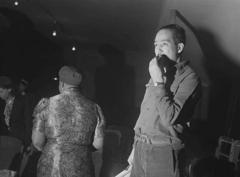 A black-and-white photo of Langston Hughes in rehearsal, taken by Jack Delano. Hughes stands in the foreground with a thoughtful expression, hand near his mouth, while another person in a patterned dress and hat is visible in the background, along with other blurred figures and dim lighting.