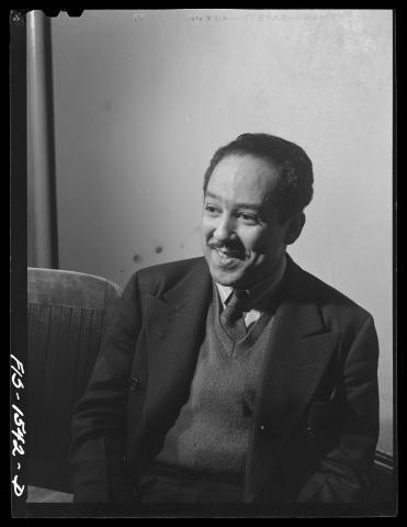A black-and-white photo of Langston Hughes, taken by Jack Delano. Hughes is seated, wearing a suit and sweater, and smiling while looking slightly to the side, with a simple backdrop and part of a chair visible.