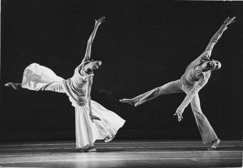 A black-and-white photo of dancers Judith Jamison and John Parks performing in Alvin Ailey's Mary Lou's Mass. They are both captured in mid-movement with arms and legs extended, Jamison wearing a flowing dress and Parks in flared pants, set against a dark background.