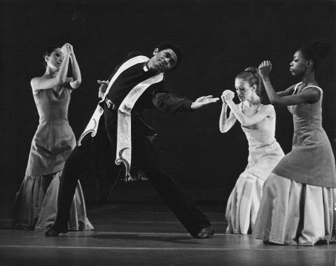 A black-and-white photo of Kelvin Rotardier in the center, performing with the Alvin Ailey American Dance Theater company in Alvin Ailey's Mary Lou's Mass. Rotardier, dressed in a priest-like robe, strikes a dramatic pose while flanked by three female dancers in long dresses, all engaged in expressive movements.