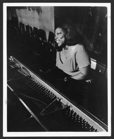 A black-and-white photo of Mary Lou Williams playing the piano, captured in a candid moment with a joyful expression. She is seated in a dimly lit room, wearing a short-sleeved top and a necklace.