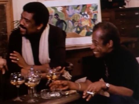 A color photo of Alvin Ailey and Romare Bearden sitting together, smiling and conversing. They are seated at a table with drinks and a colorful painting in the background.