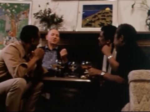 A color photo of Alvin Ailey, Romare Bearden, and two other men sitting around a table, engaged in conversation. They are in a room decorated with colorful paintings and plants, with drinks on the table in front of them.
