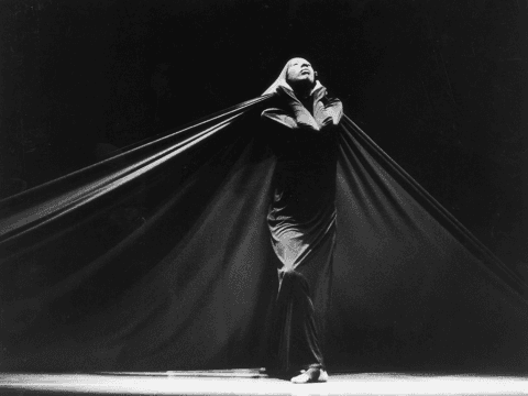 A black-and-white photo of Judith Jamison performing in Passage, enveloped in a long, flowing fabric that she holds dramatically with both hands. The background is dark, emphasizing her expressive posture and the billowing fabric.