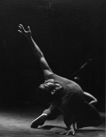 A black-and-white photo of Alvin Ailey performing a dance move from Revelations, captured in a low lunge with one arm extended upwards. The image has a dark background, emphasizing his form and movement.