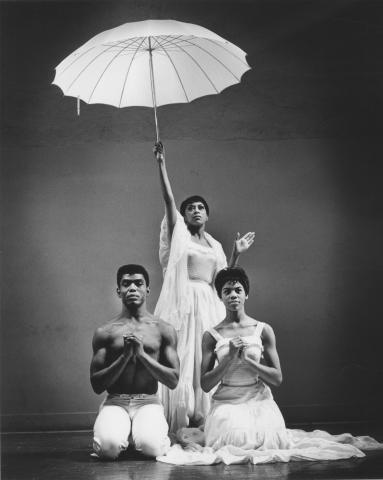 A black-and-white photo of Alvin Ailey, Ella Thompson Moore, and Myrna White in a performance of Alvin Ailey's Revelations. The image shows the three dancers in white costumes, with Moore holding a white umbrella above her head, creating a dramatic and elegant scene.