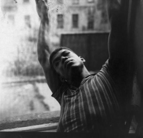 A black-and-white photo of Alvin Ailey in a striped shirt, stretching with his arms extended upwards, set against a blurred urban background. The image captures a moment of quiet intensity and focus.