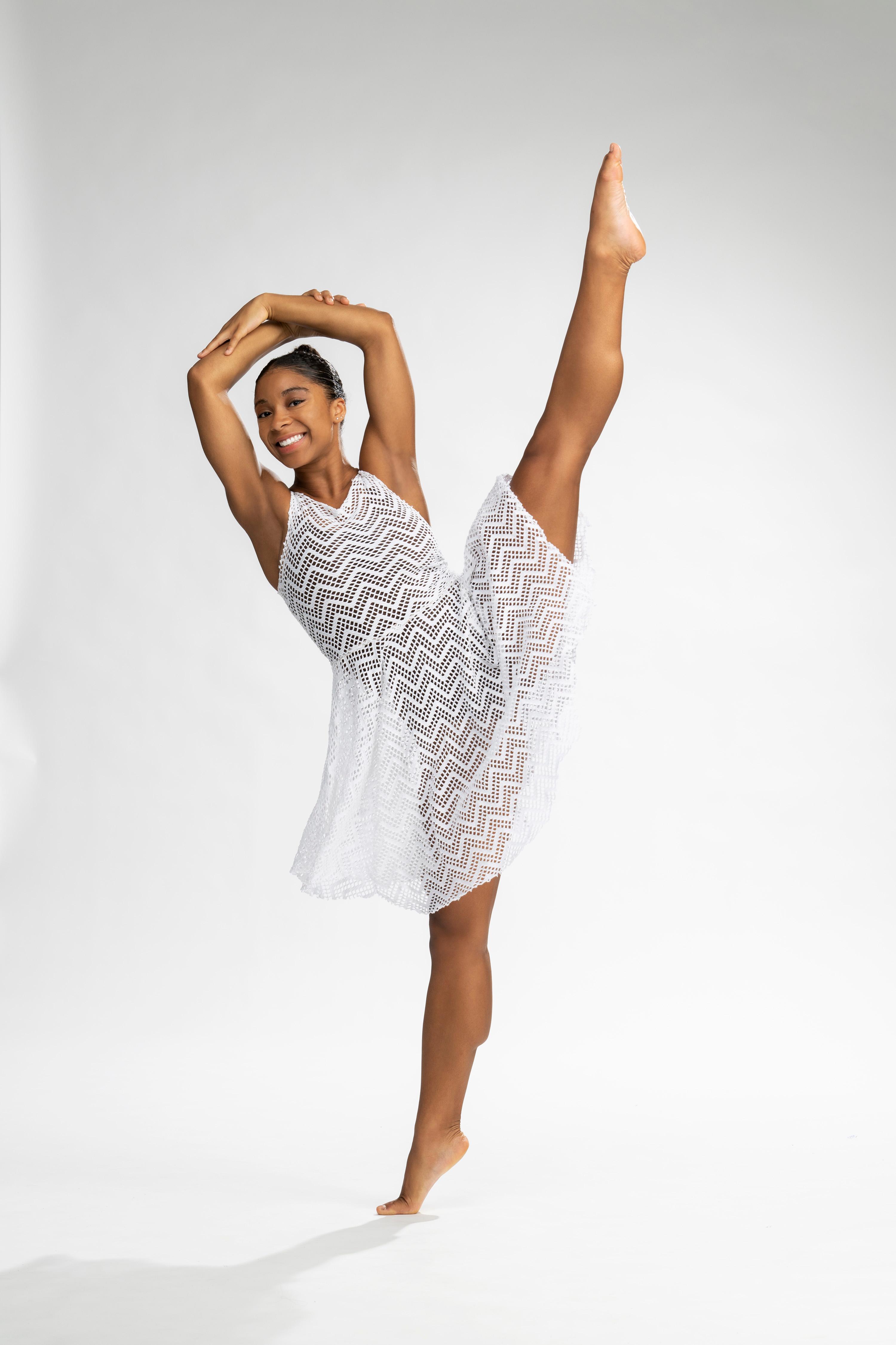 A color photo of Naia Neal, a student from the Ailey/Fordham BFA Program, performing a high leg extension while wearing a white, patterned dress. She is smiling with her arms gracefully curved above her head, set against a plain light background.