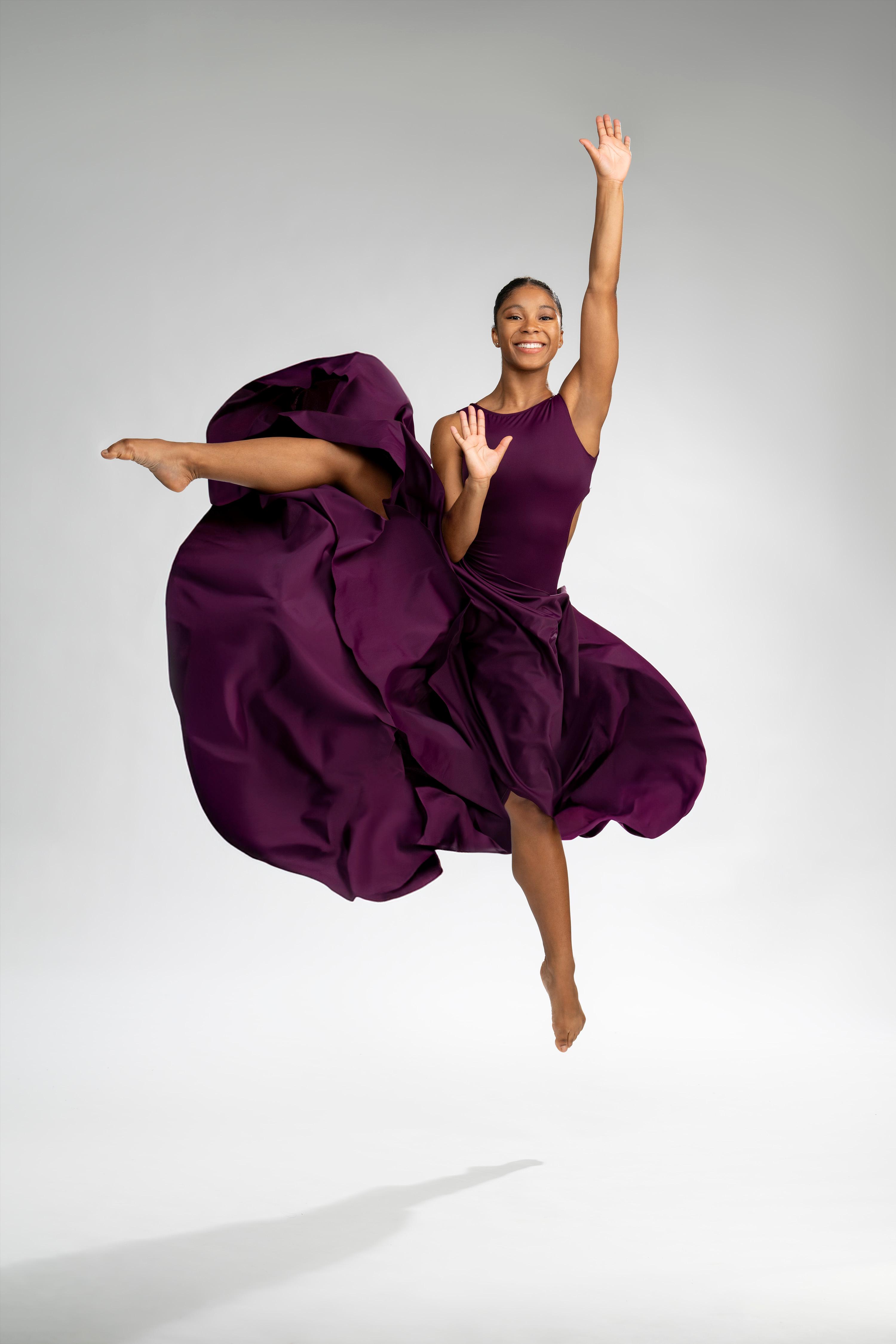A dynamic photo of Naia Neal, a student from the Ailey/Fordham BFA Program, captured mid-leap while wearing a flowing purple dress. She is smiling and extending one arm upwards and the other forward, against a plain light background.