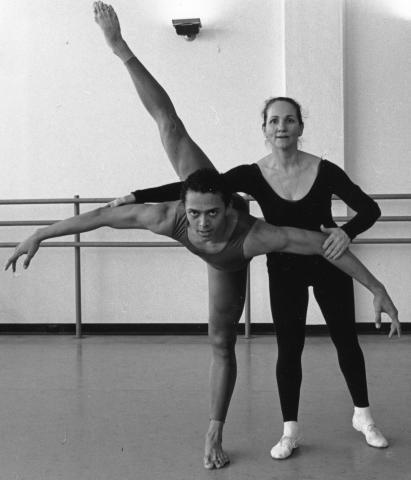Former Alvin Ailey American Dance Theater member Clifton Brown performs a high leg extension with his arms outstretched, supported by Ana Marie Forsythe, Chair of the Horton Department at The Ailey School, who stands beside him in a dance studio.