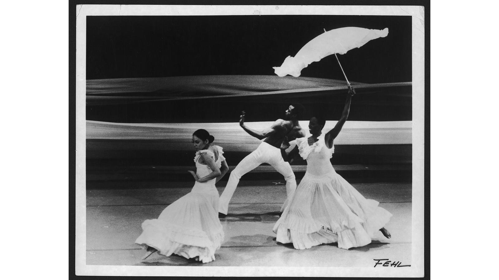 This image features dancers Mari Kajiwara, Melvin Jones, and Judith Jamison performing in Alvin Ailey’s iconic piece "Revelations." The photograph, captured by Fred Fehl, showcases the dancers in dramatic poses, with Judith Jamison holding a parasol, an element characteristic of the "Wade in the Water" section of the performance. The dancers are dressed in white, flowing costumes that add to the visual impact of the movement and the overall aesthetic of the piece. "Revelations," one of Ailey's most famous w