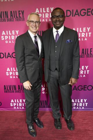 Ailey Executive Director Bennett Rink (left) and Artistic Director Robert Battle (right) stand together, smiling, in front of a backdrop featuring "Alvin Ailey American Dance Theater" and "Ailey Spirit Gala" logos. Both are dressed in suits, with Bennett Rink in a light gray suit and Robert Battle in a dark gray suit with a purple tie and pocket square.