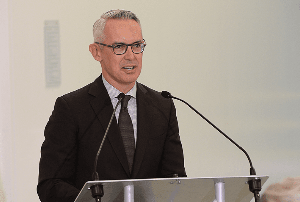 Ailey Executive Director Bennett Rink speaks at a podium with a microphone, wearing a dark suit and glasses. He is addressing an audience in a well-lit indoor setting.