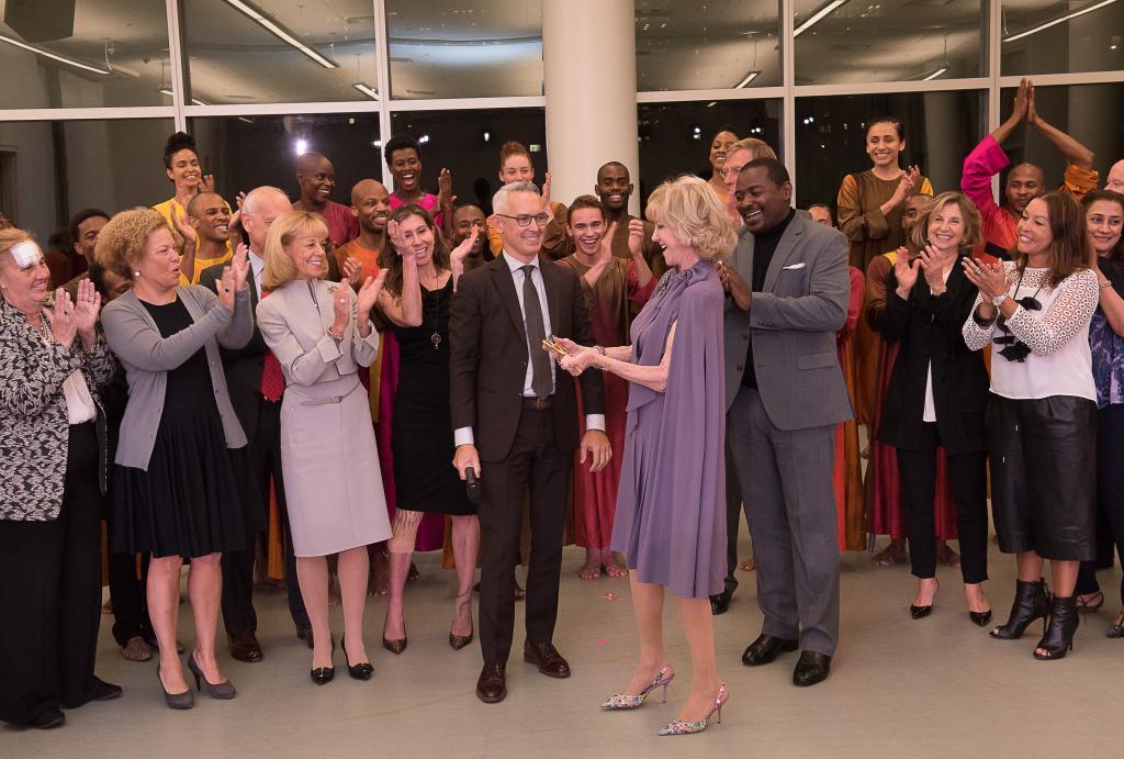 Ailey Executive Director Bennett Rink, Elaine Wynn, Artistic Director Robert Battle, and a group of board members, government officials, building architects, and Alvin Ailey American Dance Theater dancers gather for a celebration, with many clapping and smiling. The event takes place in a brightly lit room with large windows in the background.