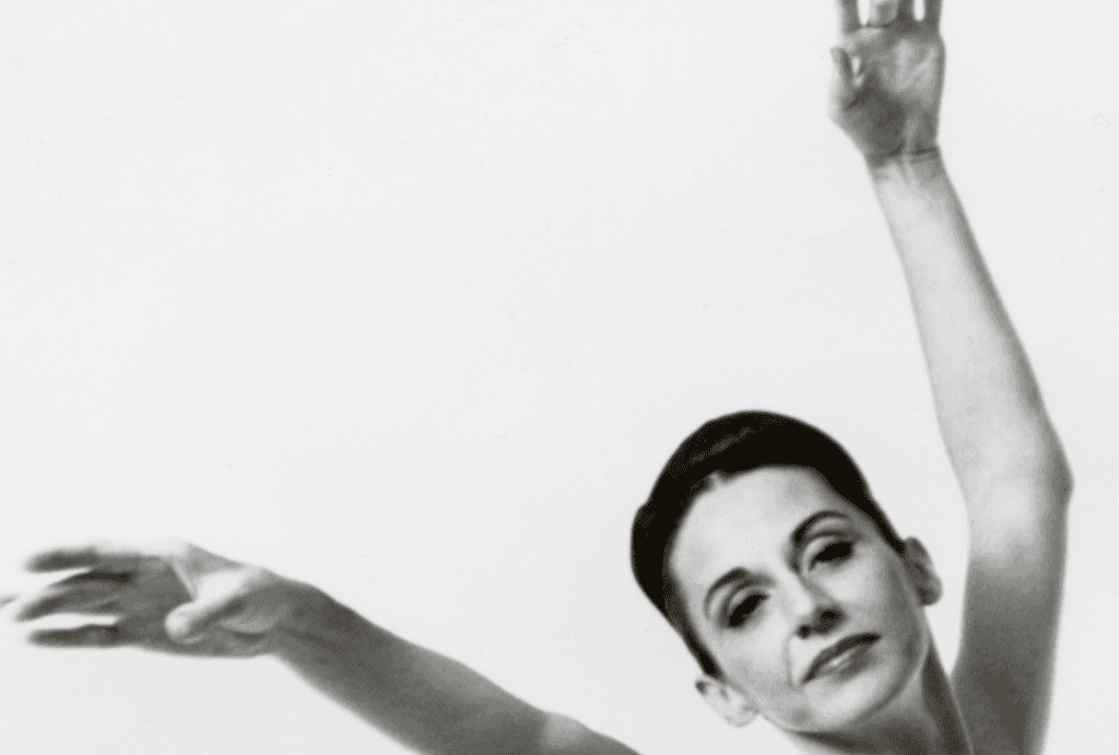 This elegant black-and-white photograph features Joyce Trisler, a prominent figure in dance. Captured in a graceful pose, Trisler's expression is serene, reflecting her deep connection to the art form. Her arms are extended delicately, showcasing her poise and control. The simplicity of the background ensures that all attention is on Trisler, emphasizing her elegance and the fluidity of her movements.