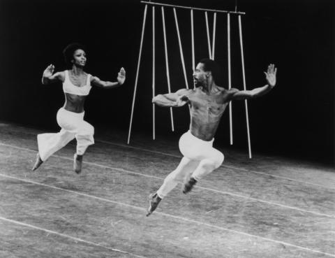 A black-and-white photo of Sylvia Waters and Dudley Williams performing in Alvin Ailey's Metallics. They are both captured mid-air in dynamic, expressive poses, wearing white costumes, with a minimalist set design in the background featuring vertical rods.