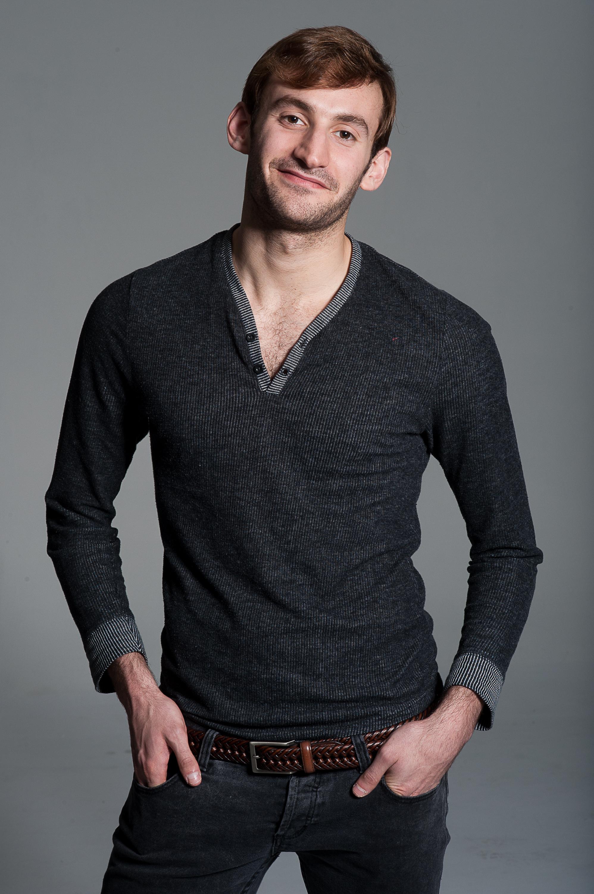 A man is smiling in his headshot. He is wearing a black V neck long sleeve top and jeans with a belt. His hands are in his pockets.
