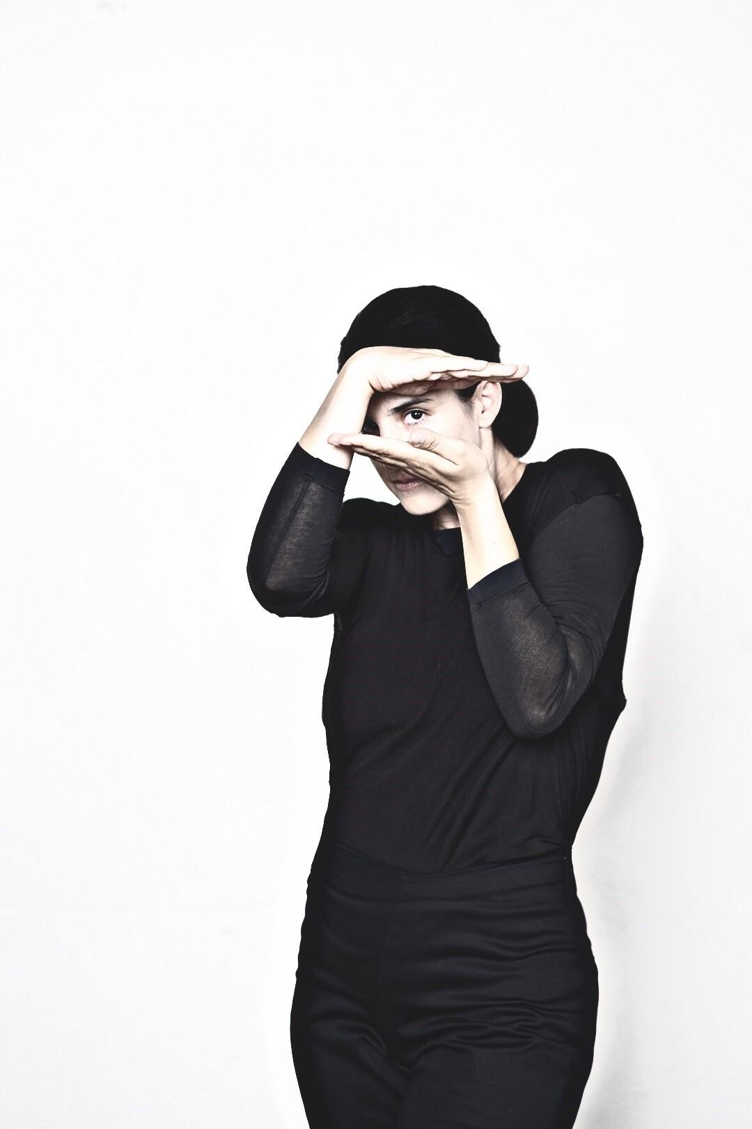 A woman is in all black with a white background. She is framing her face like she's taking a picture.