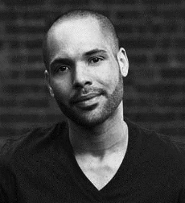 A headshot of a man in a black v neck shirt. His head is slightly tilted.