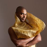 A black man with his arms folded. He has a silk yellow scarf flowing around his neck.