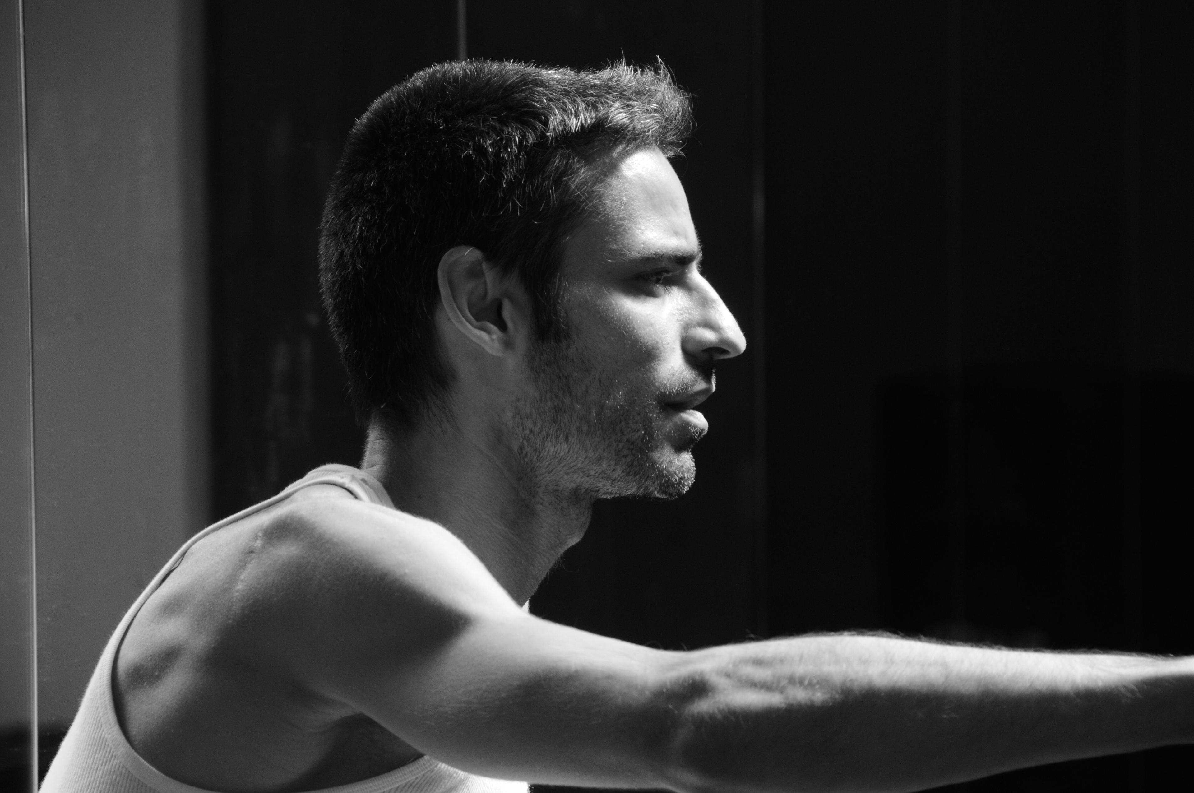 A man looks right with his arm outstretched. He is wearing a white tank. The photo is in black & white.