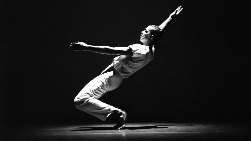 A man is facing the left in a dance pose where his knees are bent and he is leaning back. His right hand is in the air. He's in white pants and the photo is in black and white.