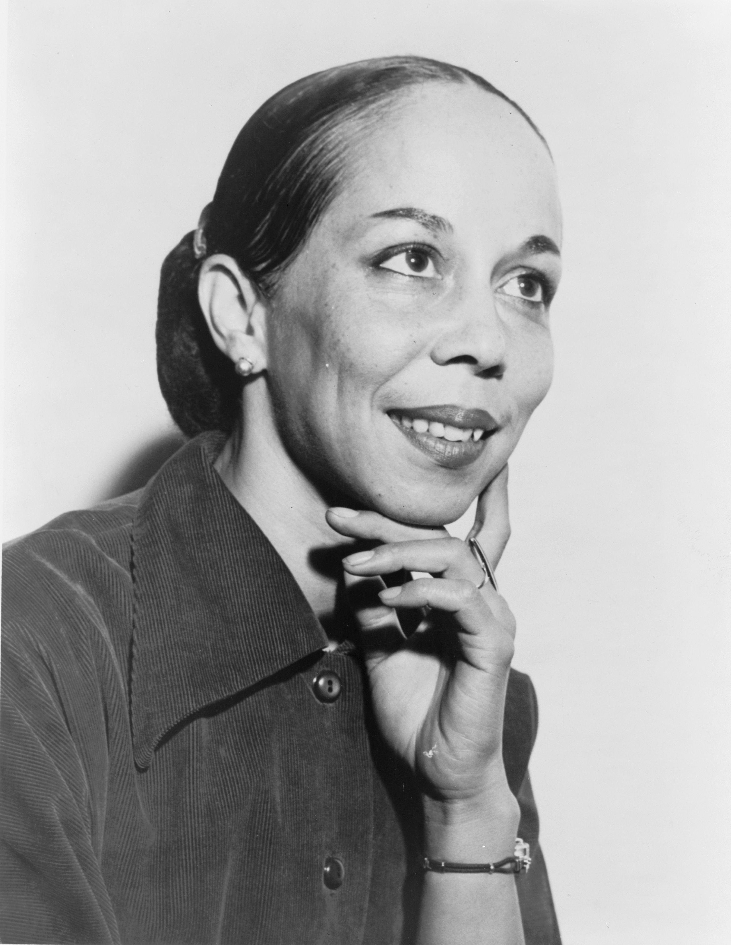 A woman is looking off to the right with her hand on her chin. Her hair is in a bun and she's wearing a collared shirt.