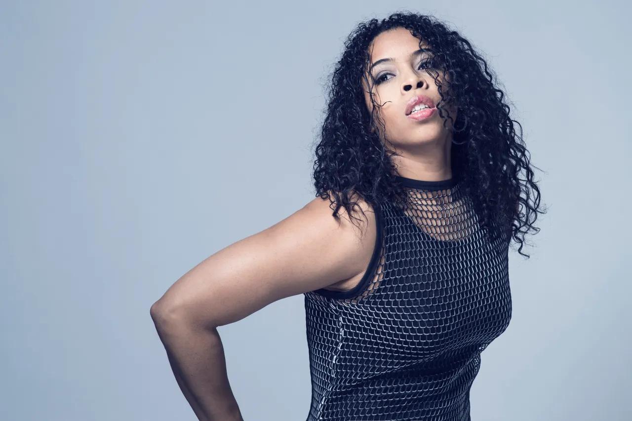 A woman poses with her hands on her hips. She is wearing a blue top with a sheer neck.