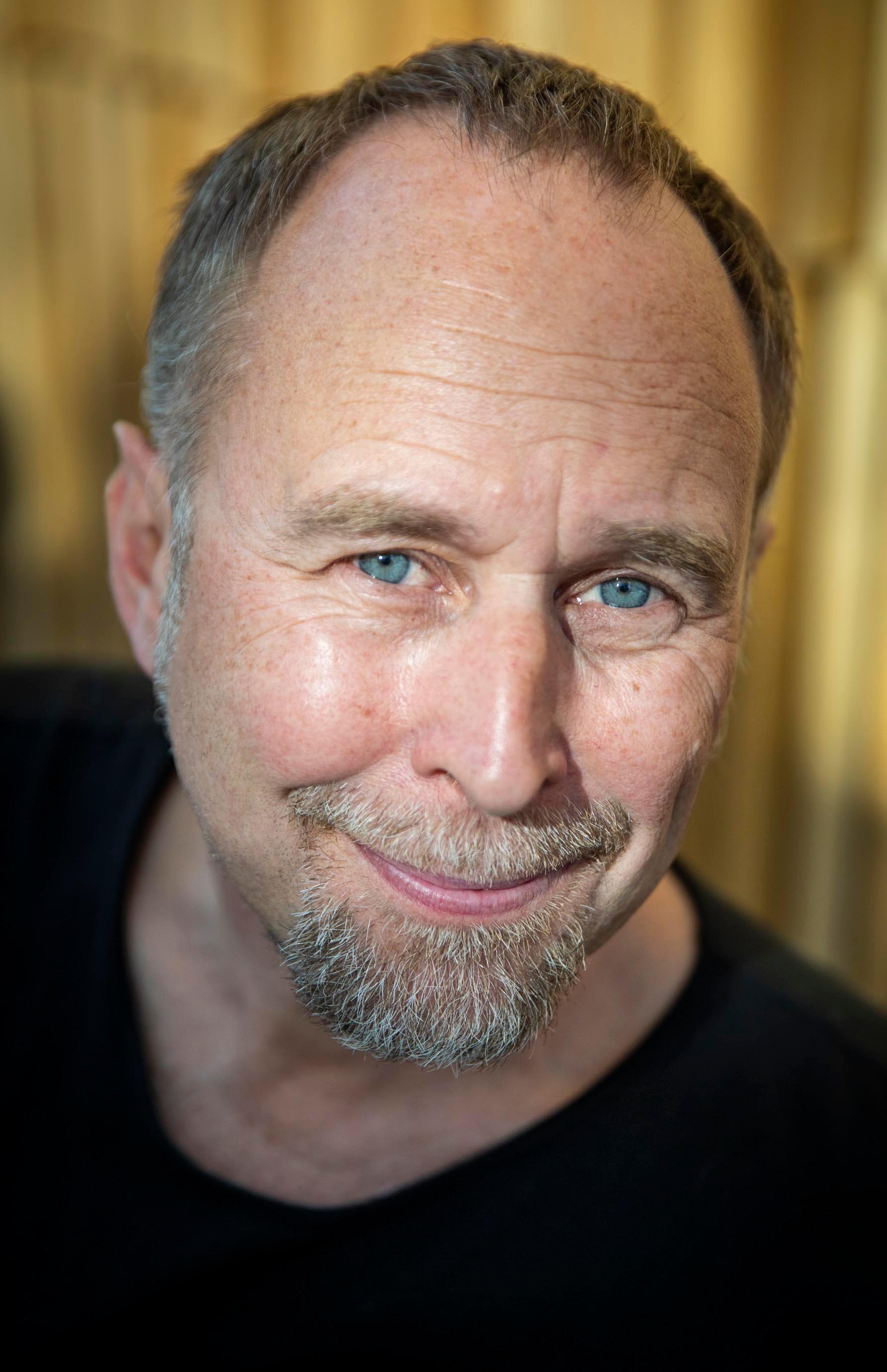 A headshot of a man with a goatee. There are yellow curtains behind him. He is wearing a black tee.
