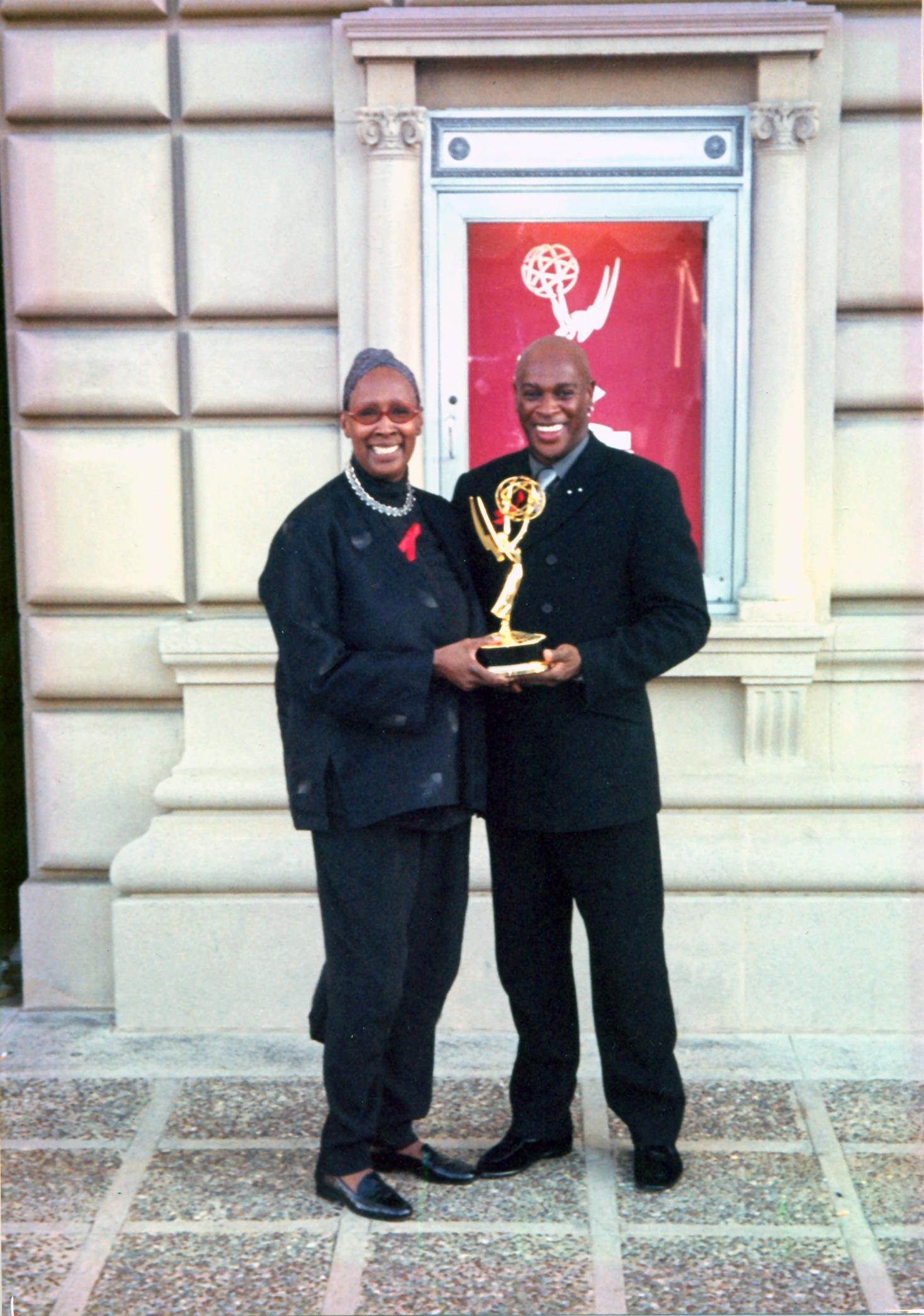 Judith Jamison and Marc Manigault holding an Emmy award outside a building. Both are smiling and dressed in dark attire. The background features a red poster with the Emmy logo. The photo is from 1999.