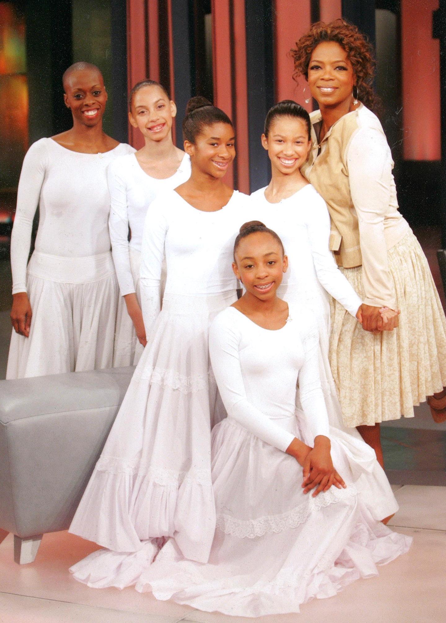 This image features students from The Ailey School along with D. Smallwood from the Alvin Ailey American Dance Theater, posing with Oprah Winfrey during a taping of "The Oprah Winfrey Show" in 2007. The students and dancer are dressed in white, reminiscent of the costumes often seen in performances of "Revelations," one of Alvin Ailey's most iconic works. Oprah Winfrey, a well-known supporter of the arts, is seen smiling and holding hands with one of the students, highlighting the connection and support be