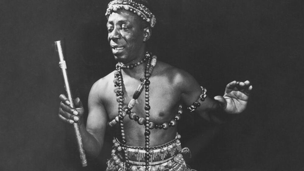 A black man is in a grass skirt with necklaces and a headband. He holds a musical instrument and seems to be dancing. The photo is black & white