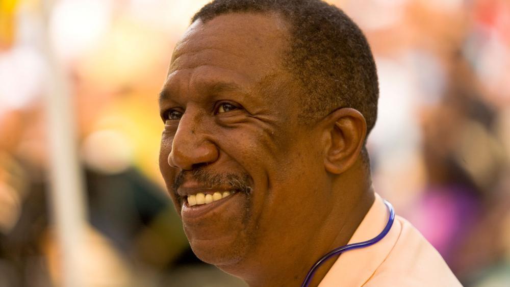 A black man smiles and looks to the left. There are blurred colors behind him. He is wearing a salmon shirt with a blue cord.