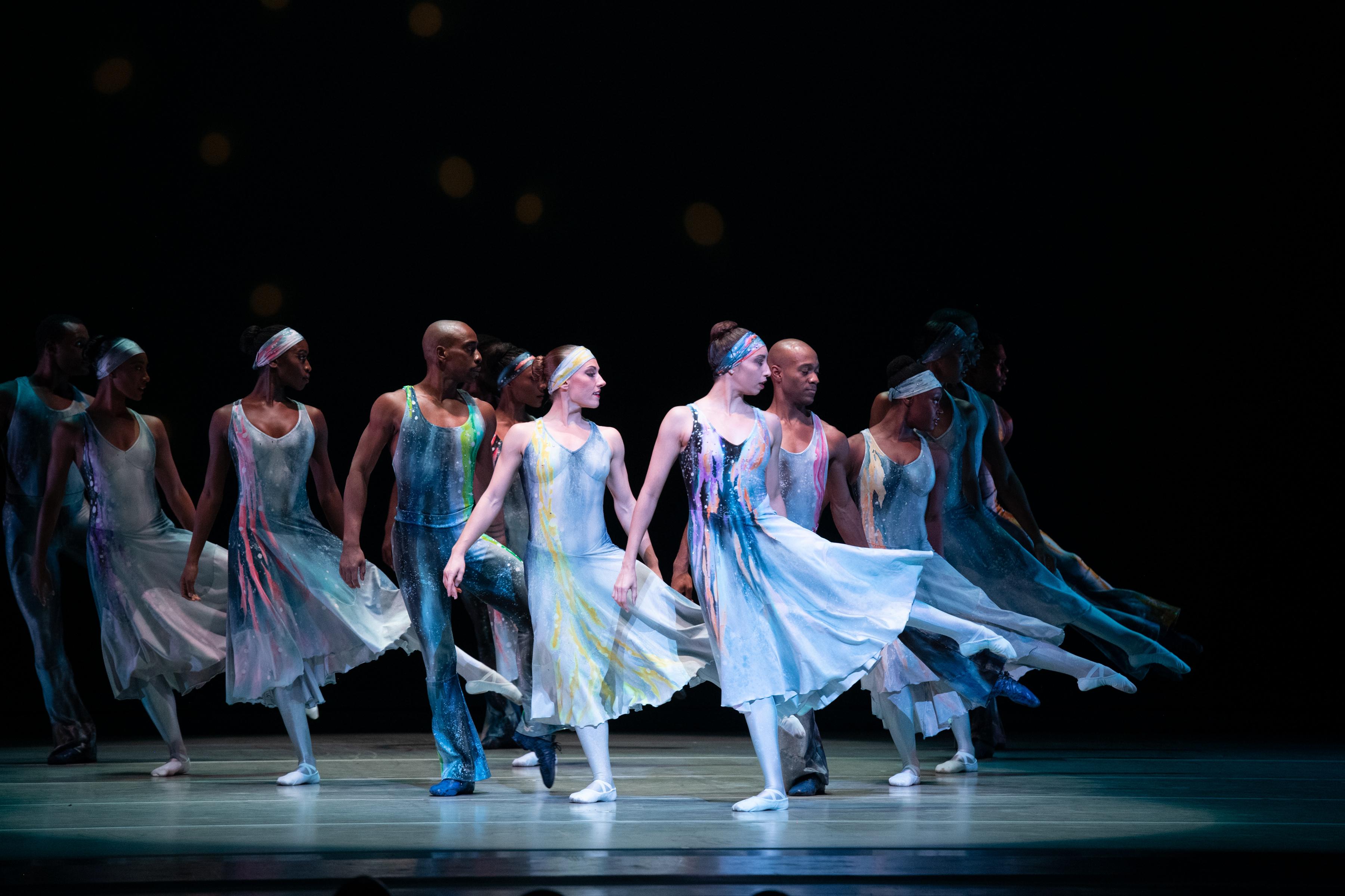 A group of dancers in blue dresses on stage. They are all looking right with their leg pointed in that direction.
