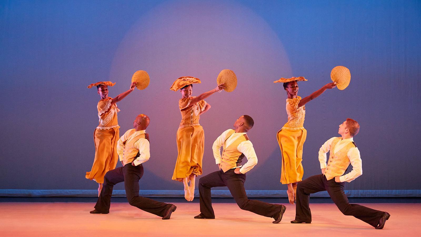 Six dancers perform on stage against a blue background. Three female dancers in yellow costumes and hats are mid-jump, holding fans. Three male dancers in white shirts and yellow vests kneel with one knee up, looking up at the female dancers. The stage is lit with warm lighting.