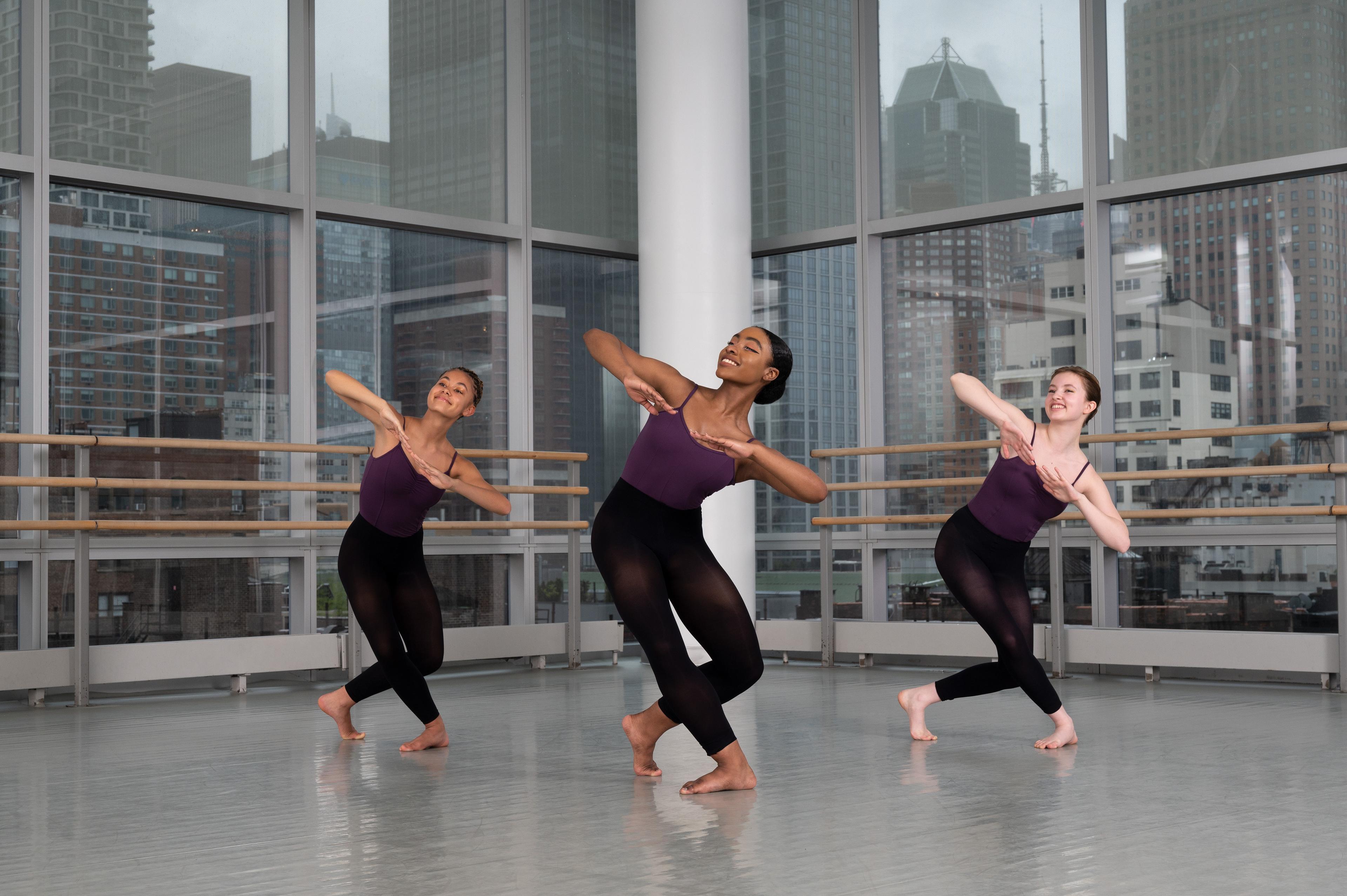 Three girls standing in a triangle on bended knees leaning to their left side and looking up. They are all wearing purple leotards with black tights over them and bare feet.