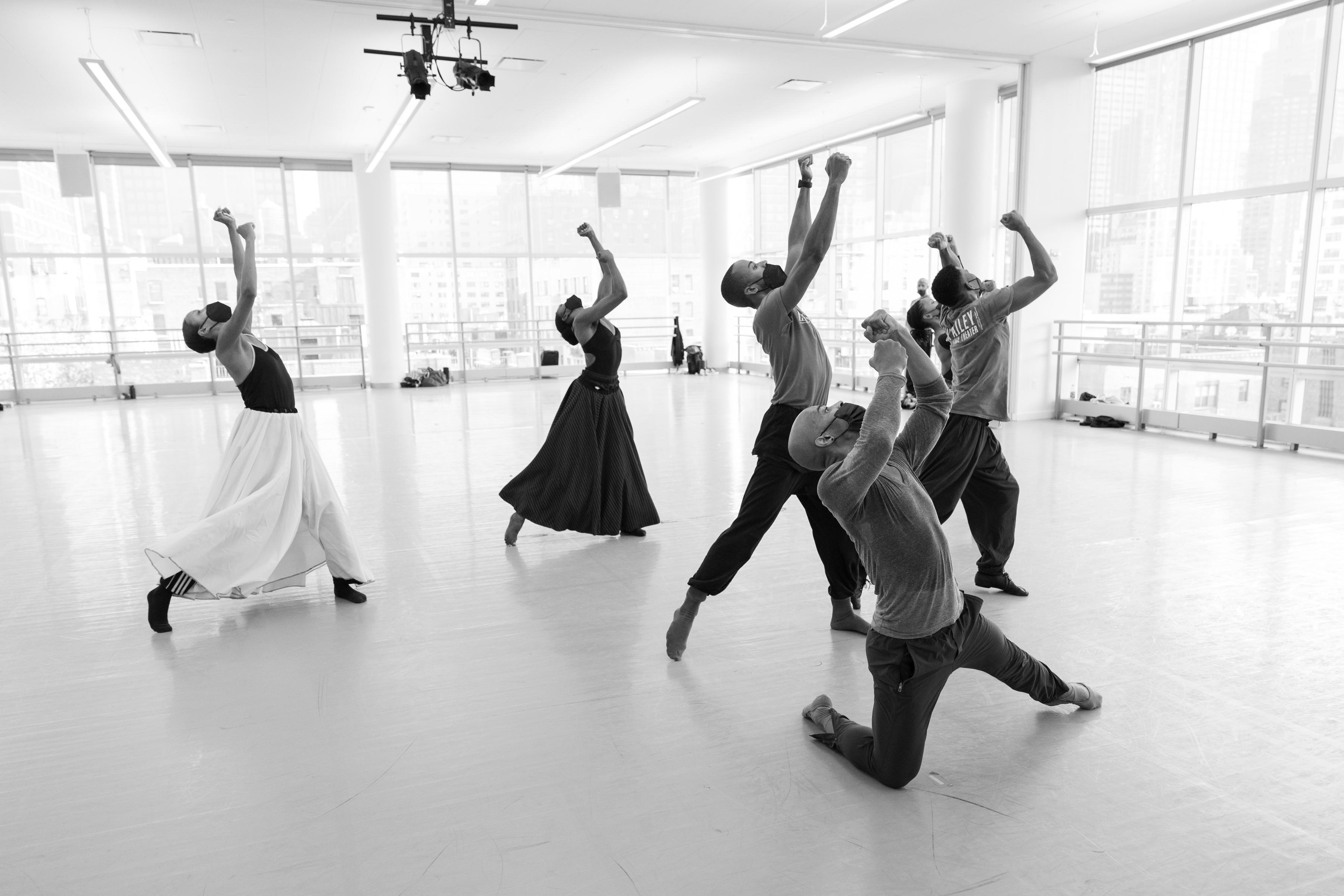 The black-and-white image captures a poignant moment during a rehearsal of Alvin Ailey's "Survivors" by the Alvin Ailey American Dance Theater. The dancers are positioned in expressive, upward-reaching postures, conveying a sense of struggle and determination. Their movements create strong, dynamic lines, emphasizing the emotional depth of the piece. The studio's expansive windows flood the room with natural light, illuminating the dancers and highlighting their powerful physicality.
