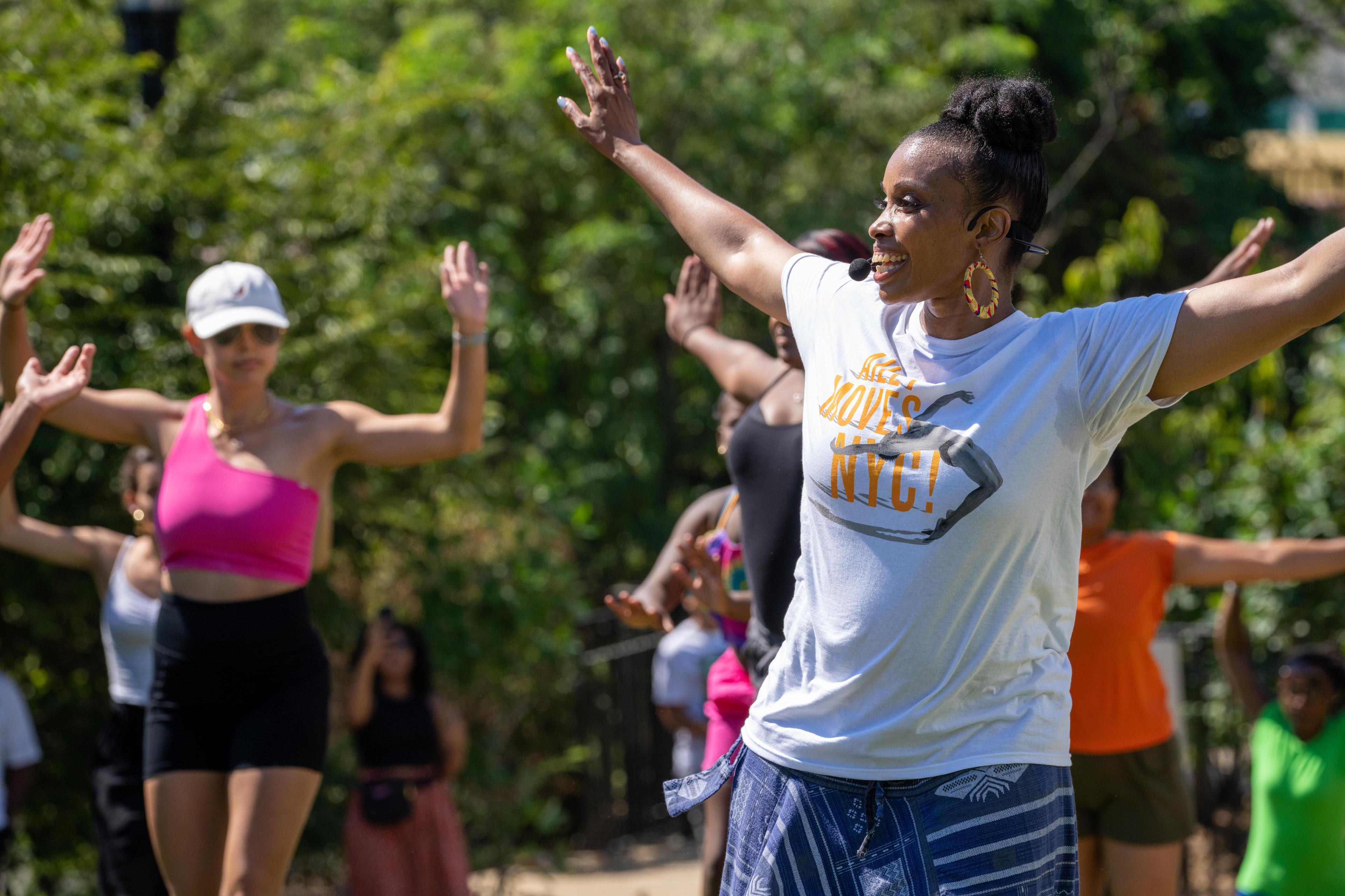 A color photo of Fatima Logan-Alston leading a West African dance workshop at Coney Island. She is wearing a white t-shirt and patterned pants, smiling with arms raised, while participants behind her follow the movements, set against a backdrop of greenery.
