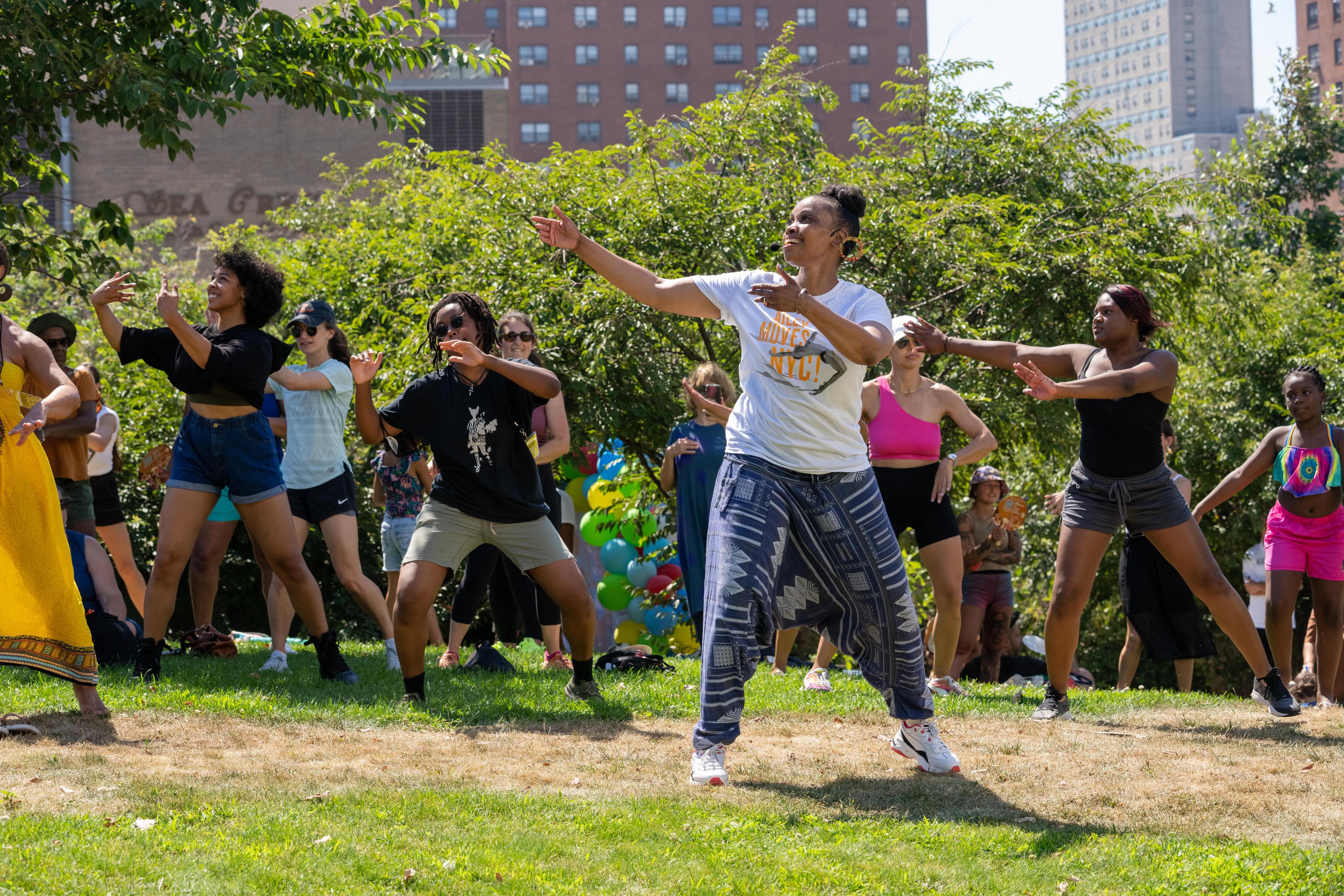 A color photo of Fatima Logan-Alston leading a West African dance workshop at Coney Island, surrounded by a diverse group of participants. They are dancing outdoors on a grassy area with trees and buildings in the background, under a sunny sky.